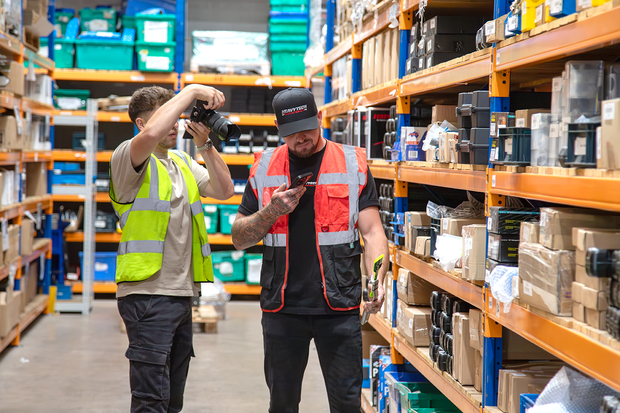 Two people in the warehouse, one taking a photo with a smartphone, the other holding a camera.